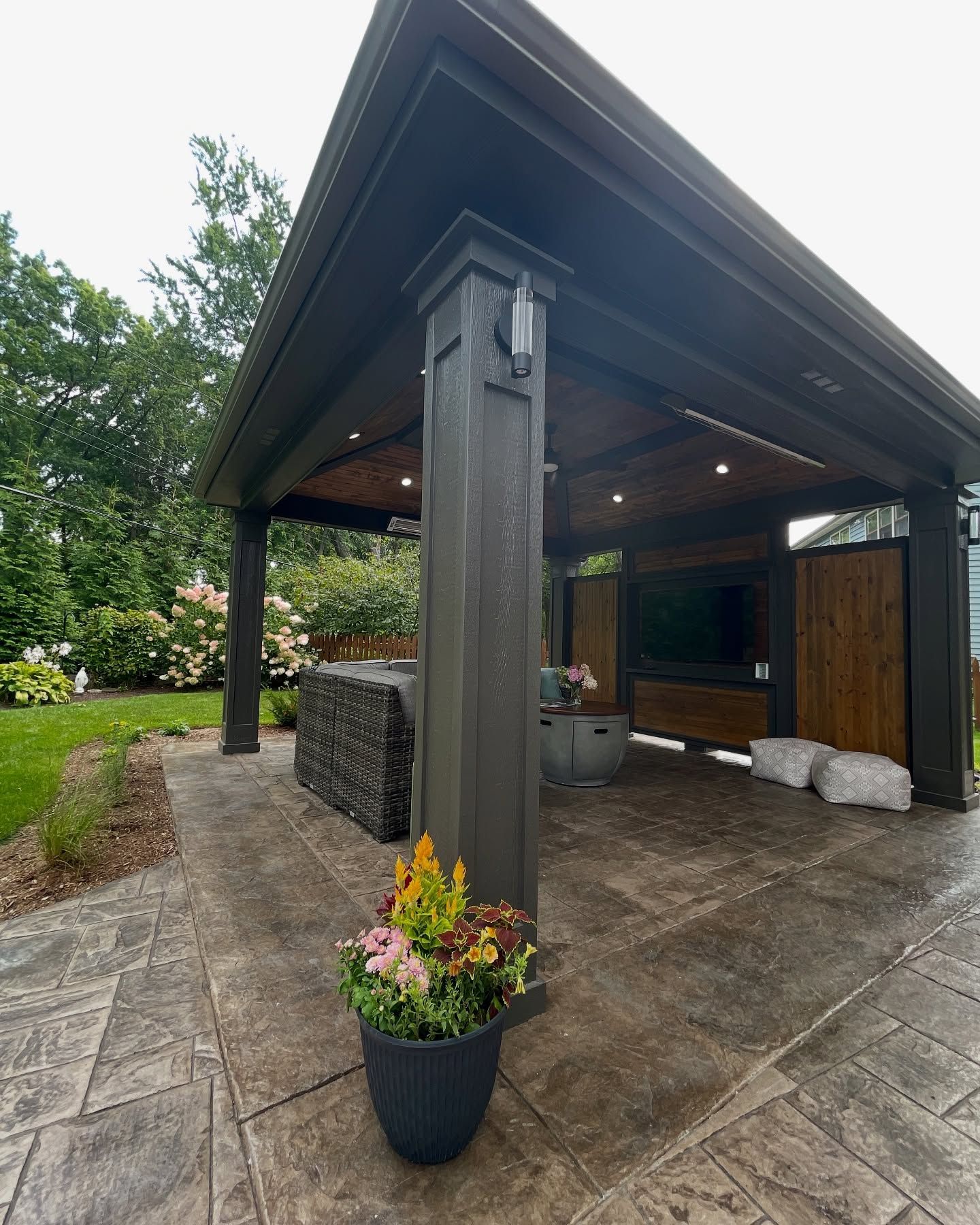 Outdoor covered patio with a large-screen TV and bar area; dark gray structure with wooden accents, surrounded by greenery.