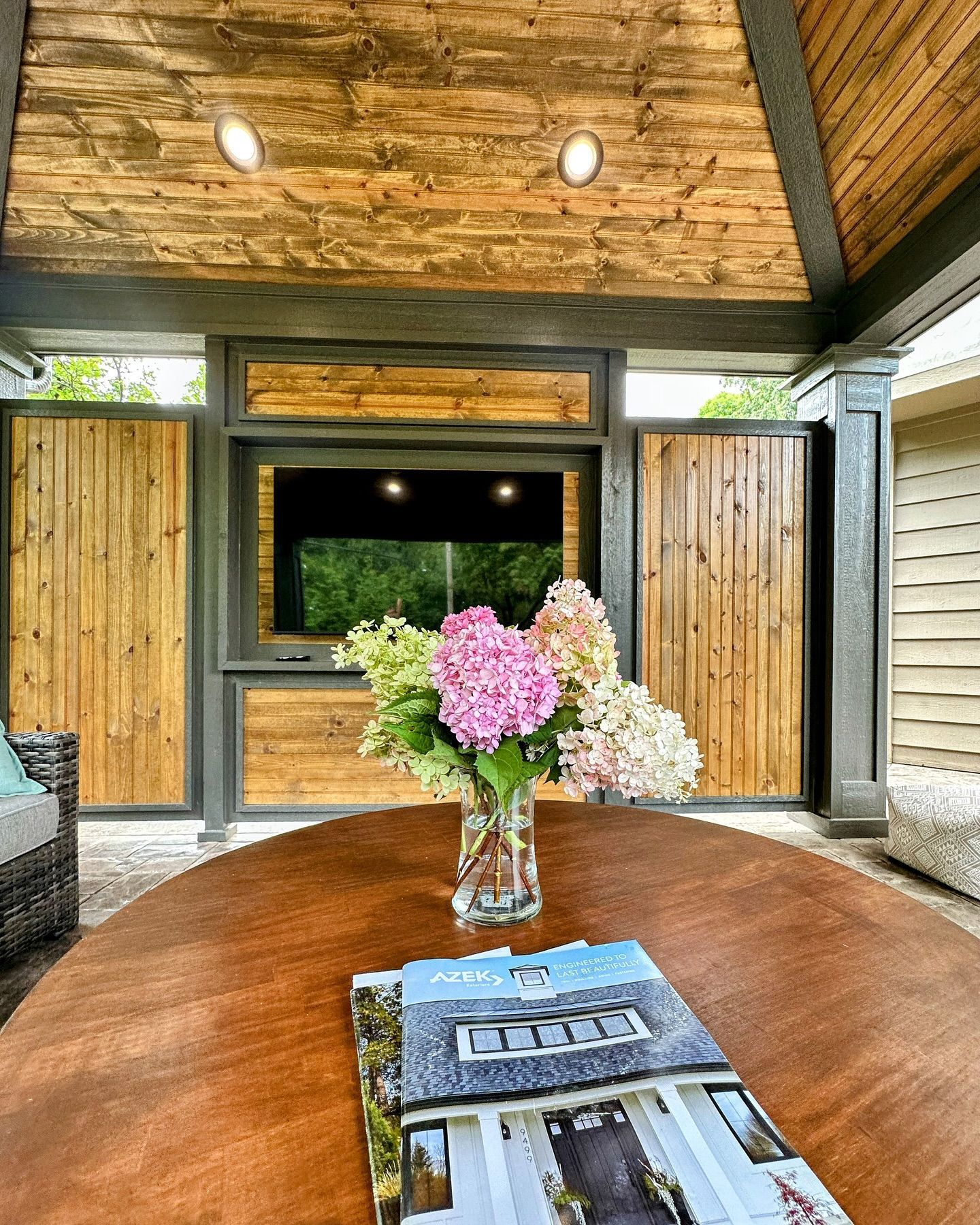 Outdoor entertainment space with wooden canopy and built-in TV, featuring a round table with flowers and a magazine.