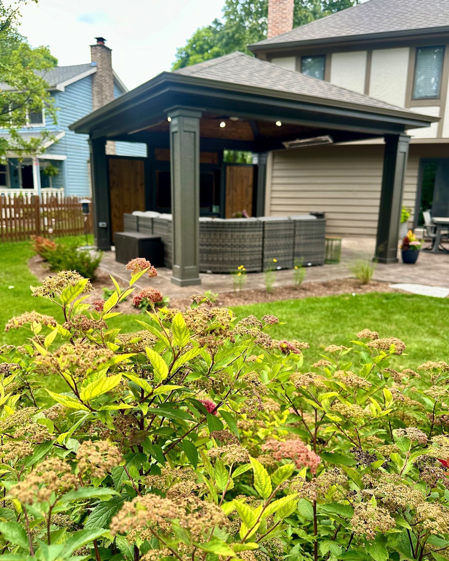 Outdoor hot tub under a gazebo, surrounded by green grass and plants. Dark brown structure, light brown house.
