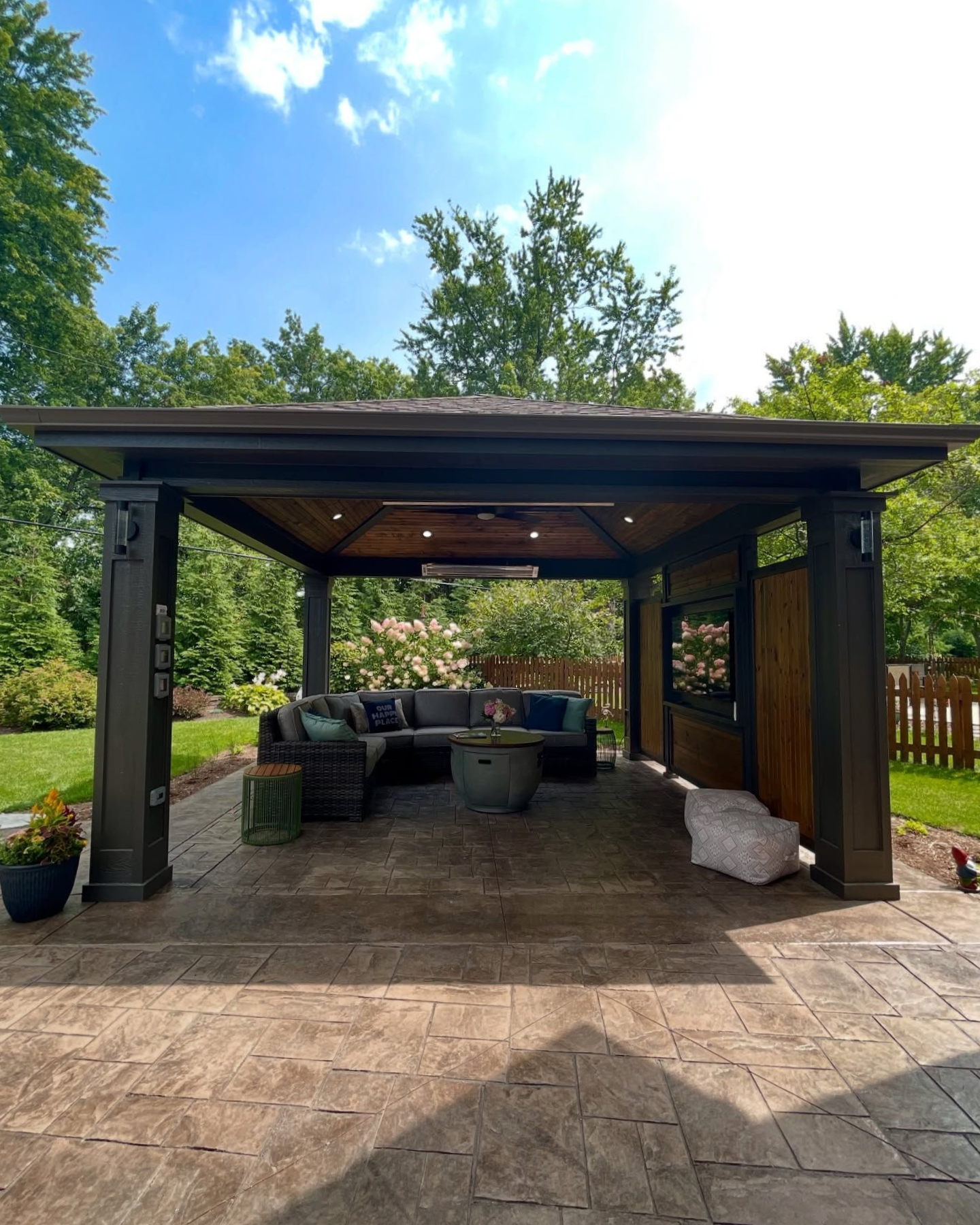 Outdoor covered patio with seating, fireplace, and green landscape under a blue sky.
