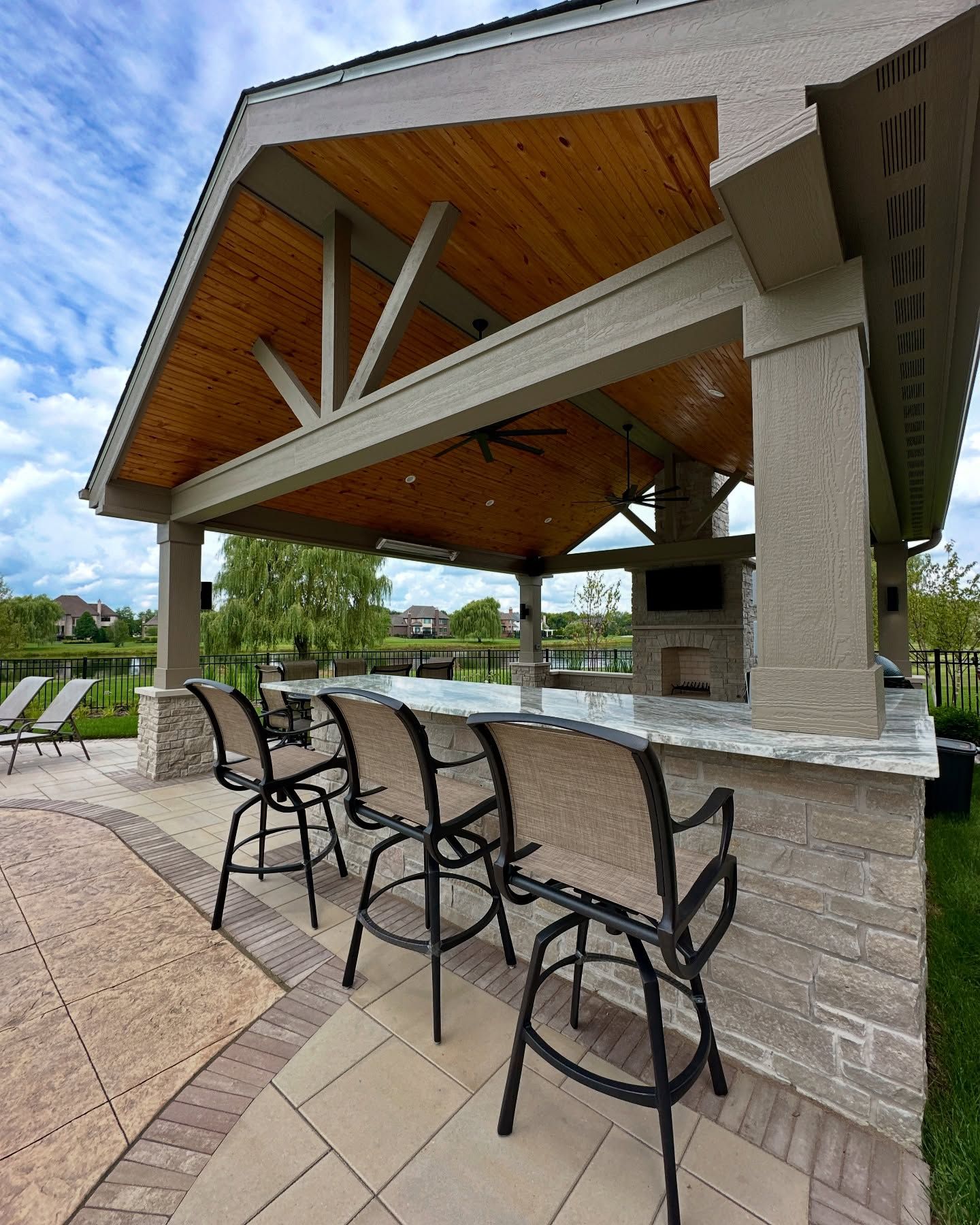 Outdoor covered bar with seating; stone, wood, and tan construction, overlooking a water view.