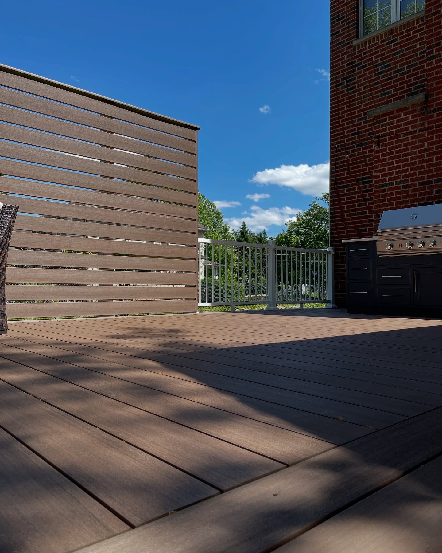 Brown composite deck with a privacy screen and brick building against a blue sky.