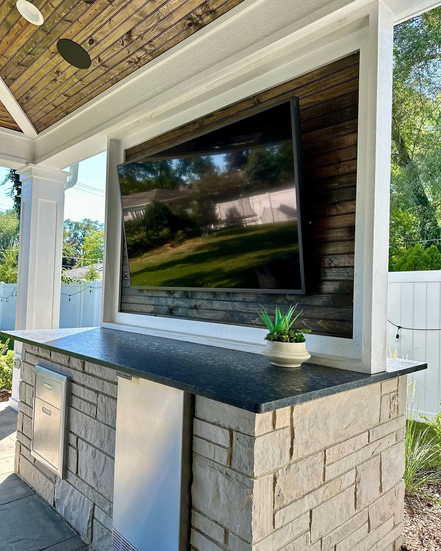 Outdoor kitchen with mounted TV, stone bar, and wooden ceiling.