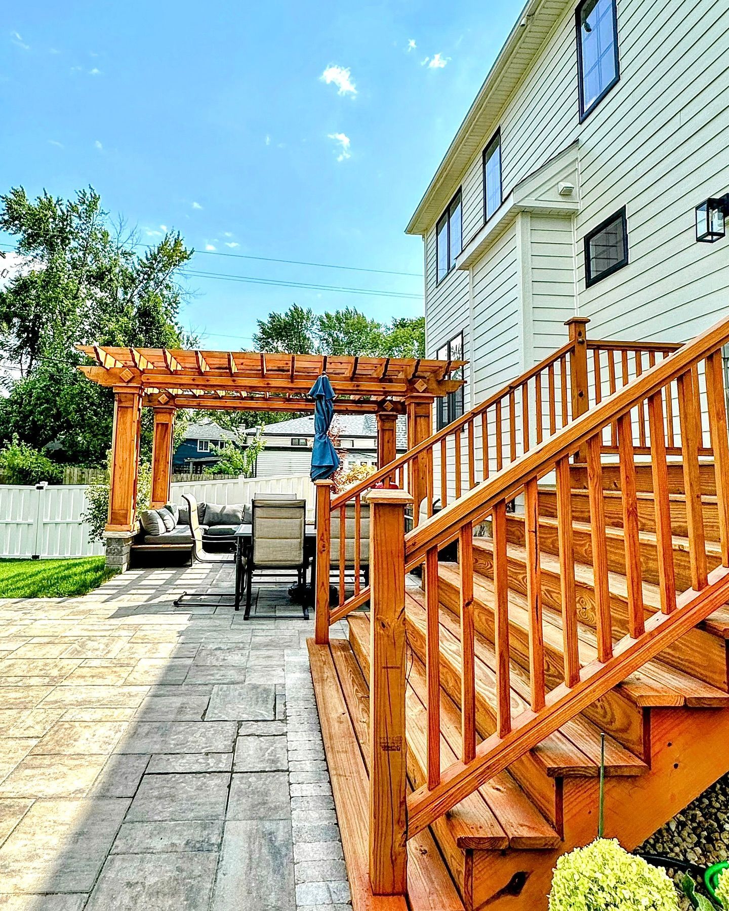 Wooden deck with steps leading to a backyard patio, pergola, seating, and a white house. Bright sunny day.