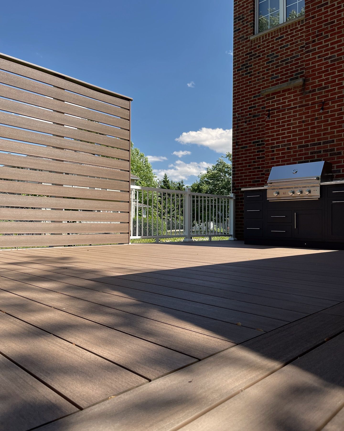 Composite deck with brown horizontal privacy screen, a brick building, and a grill under a blue sky.