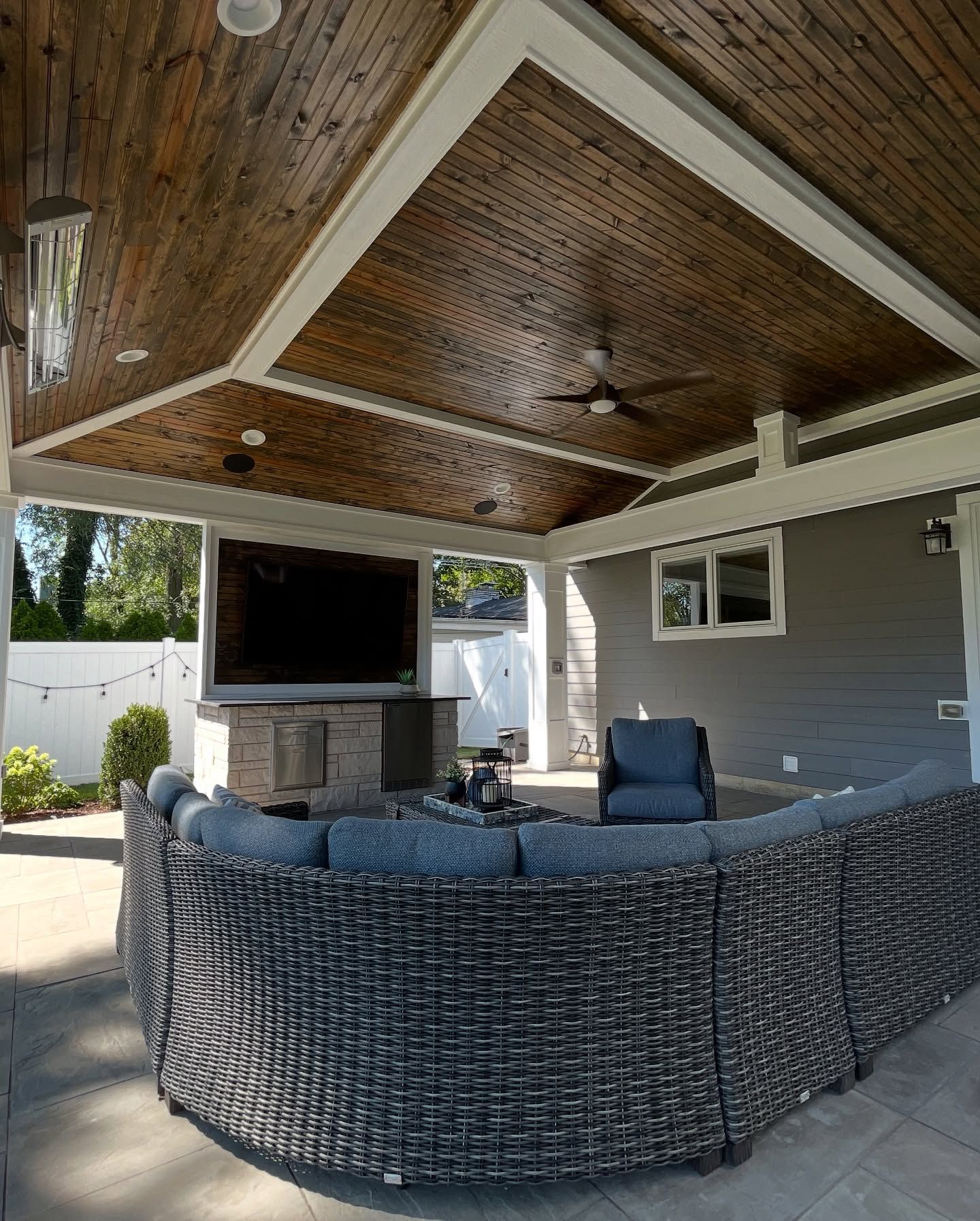 Covered patio with dark wood ceiling, white trim, outdoor TV, and curved wicker sectional.