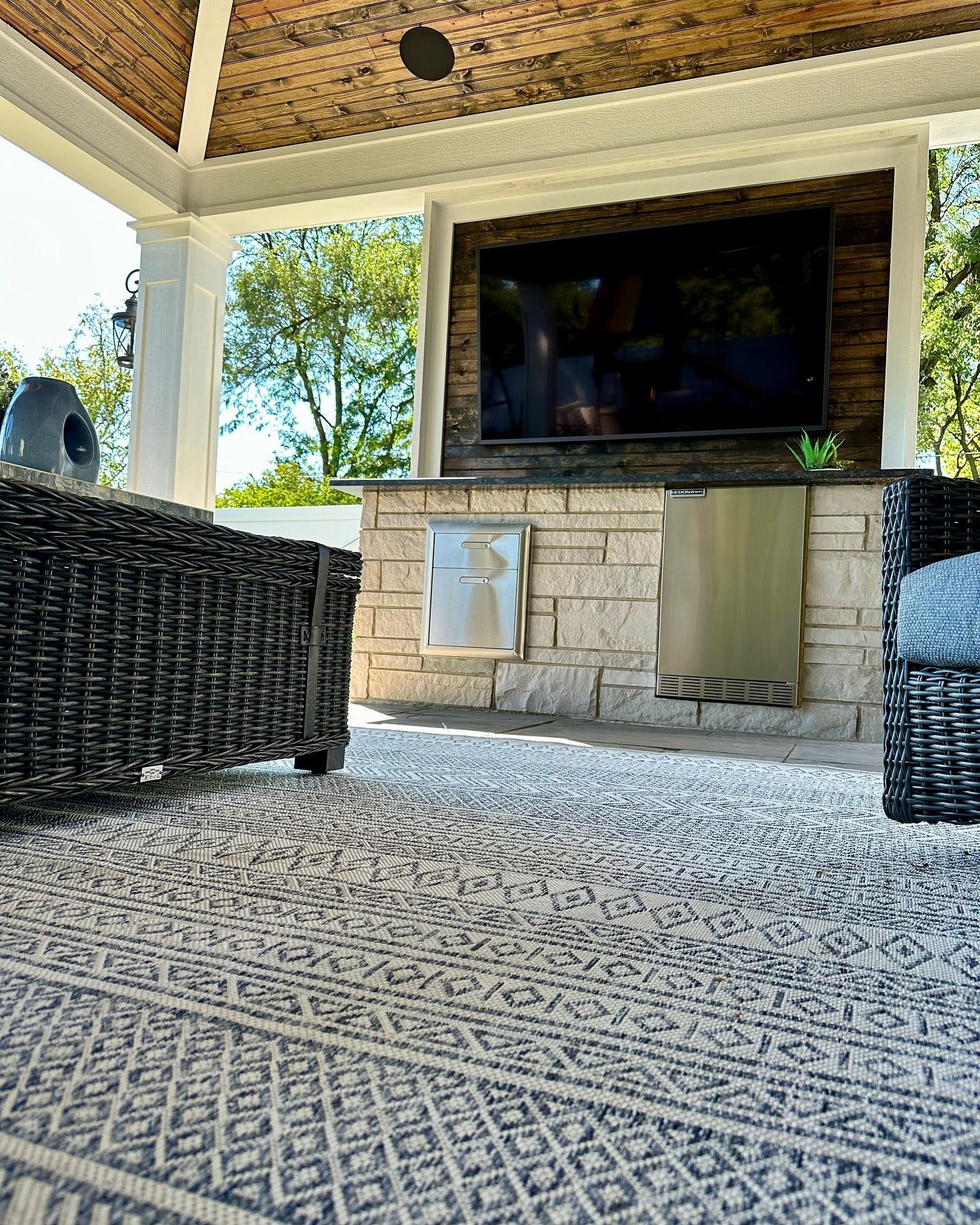 Outdoor living space with TV on stone wall, wicker furniture, and patterned rug.