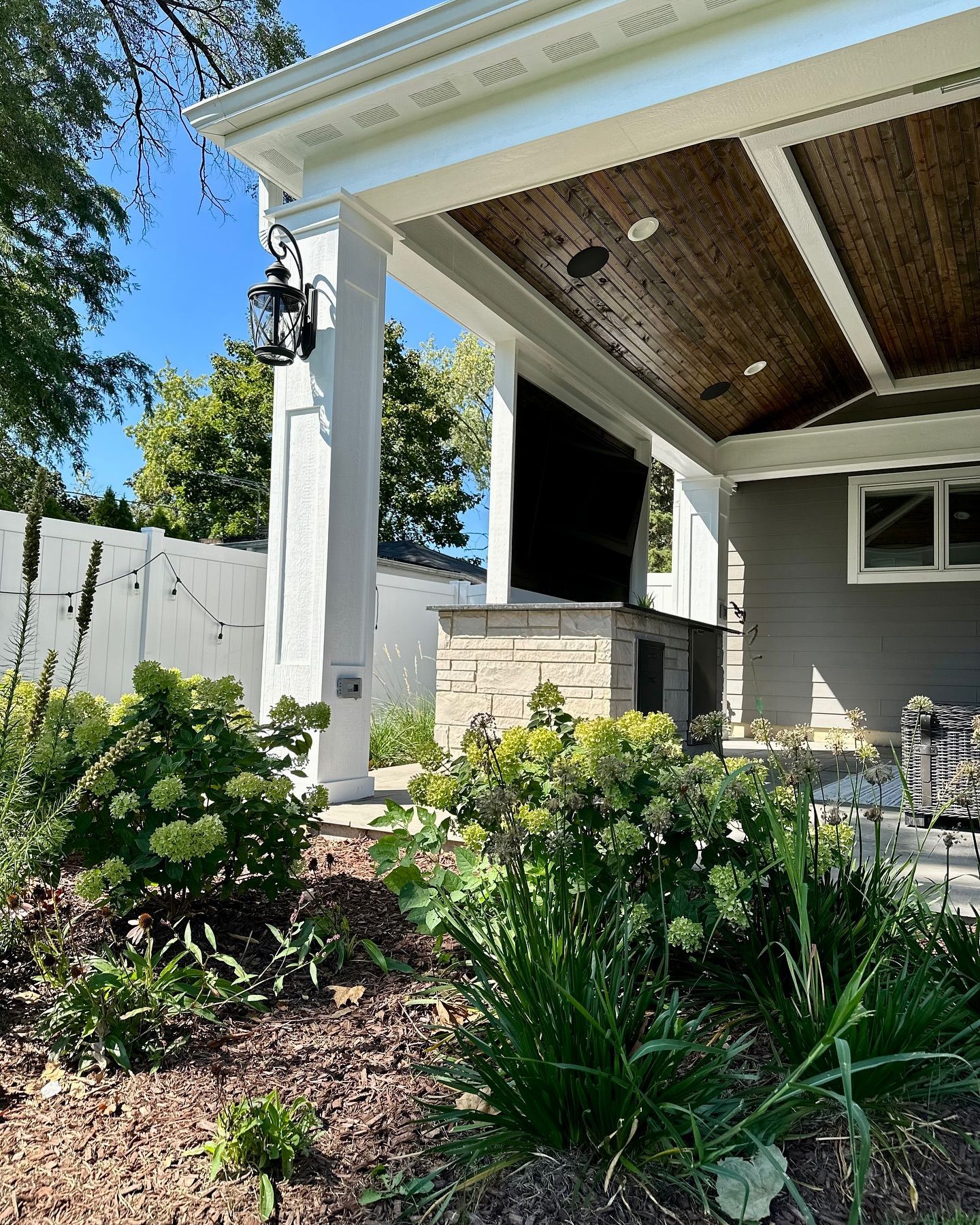 Outdoor patio with stone fireplace, TV, and garden, under a covered porch.