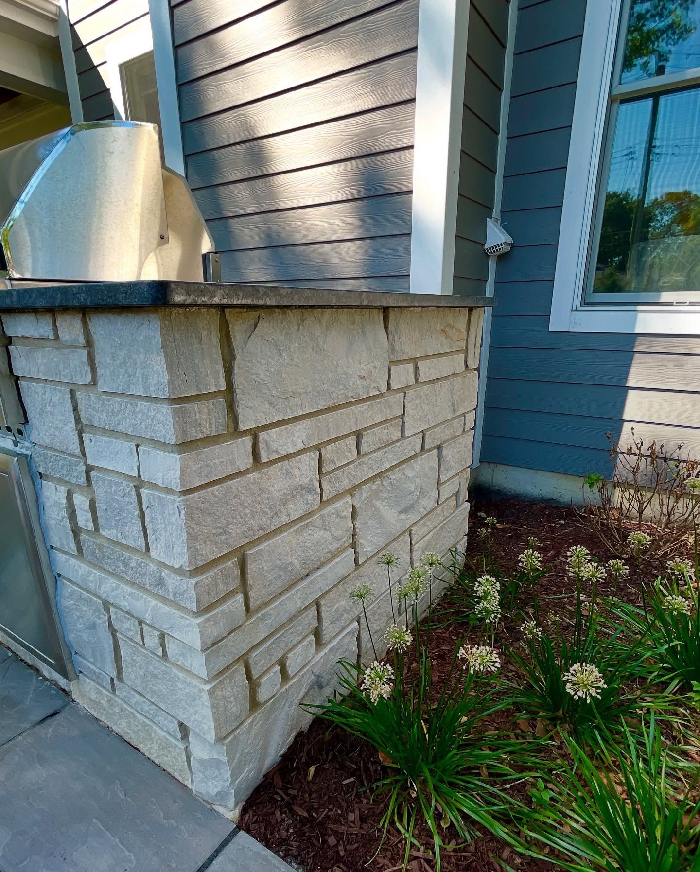Stone brick outdoor kitchen island next to a gray house, with a grill and flowers.
