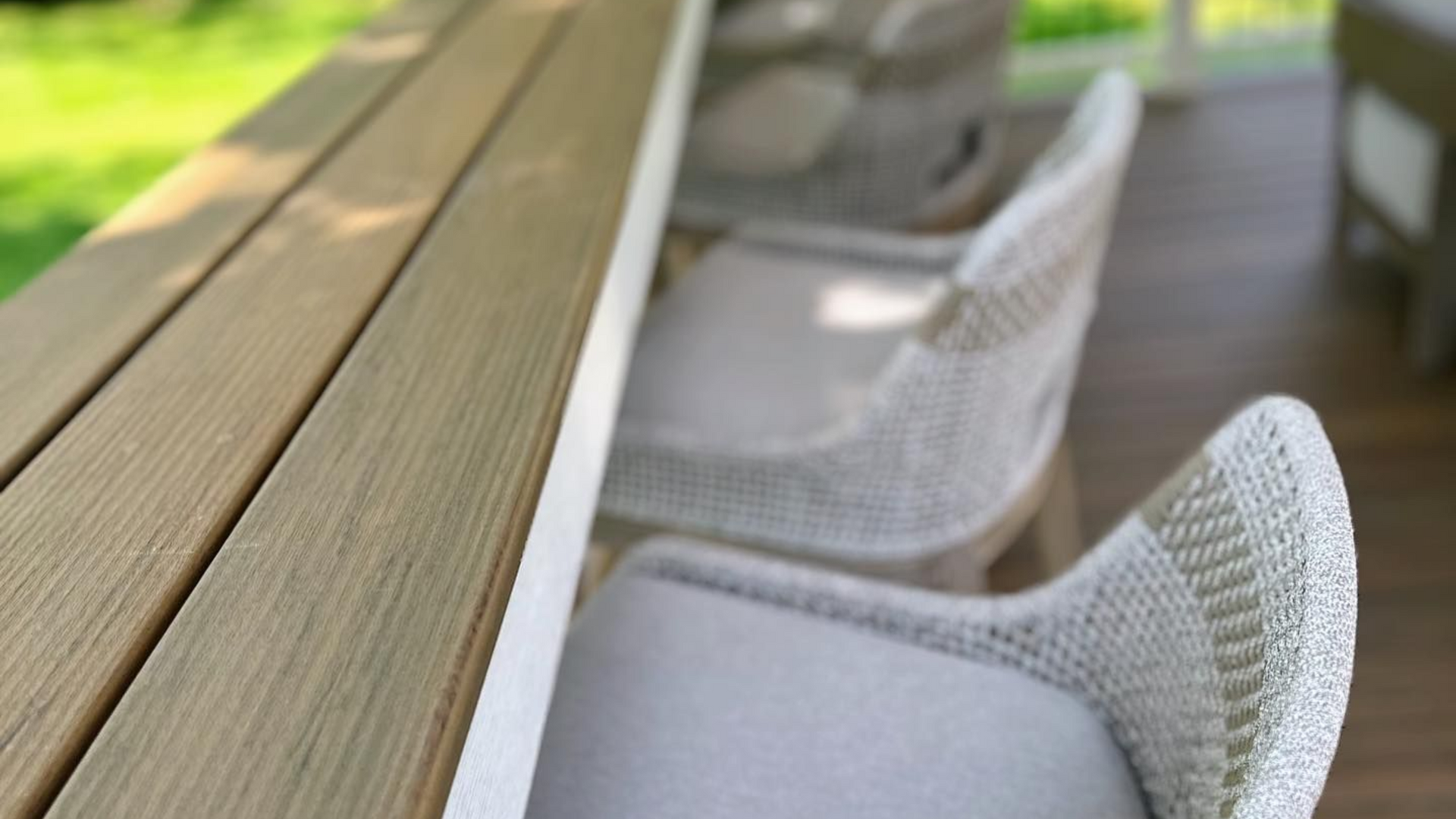 Wooden table with three woven chairs on a porch. Green foliage in the background.