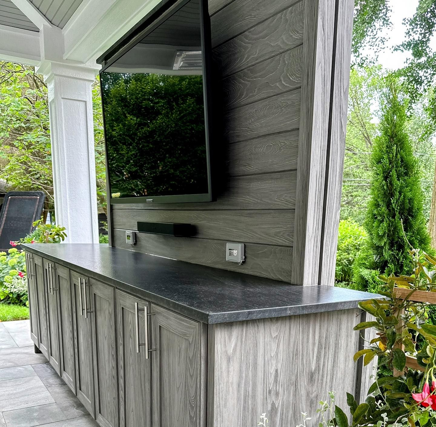 Outdoor entertainment cabinet with TV, dark granite countertop, and gray wood cabinets. Green foliage in background.