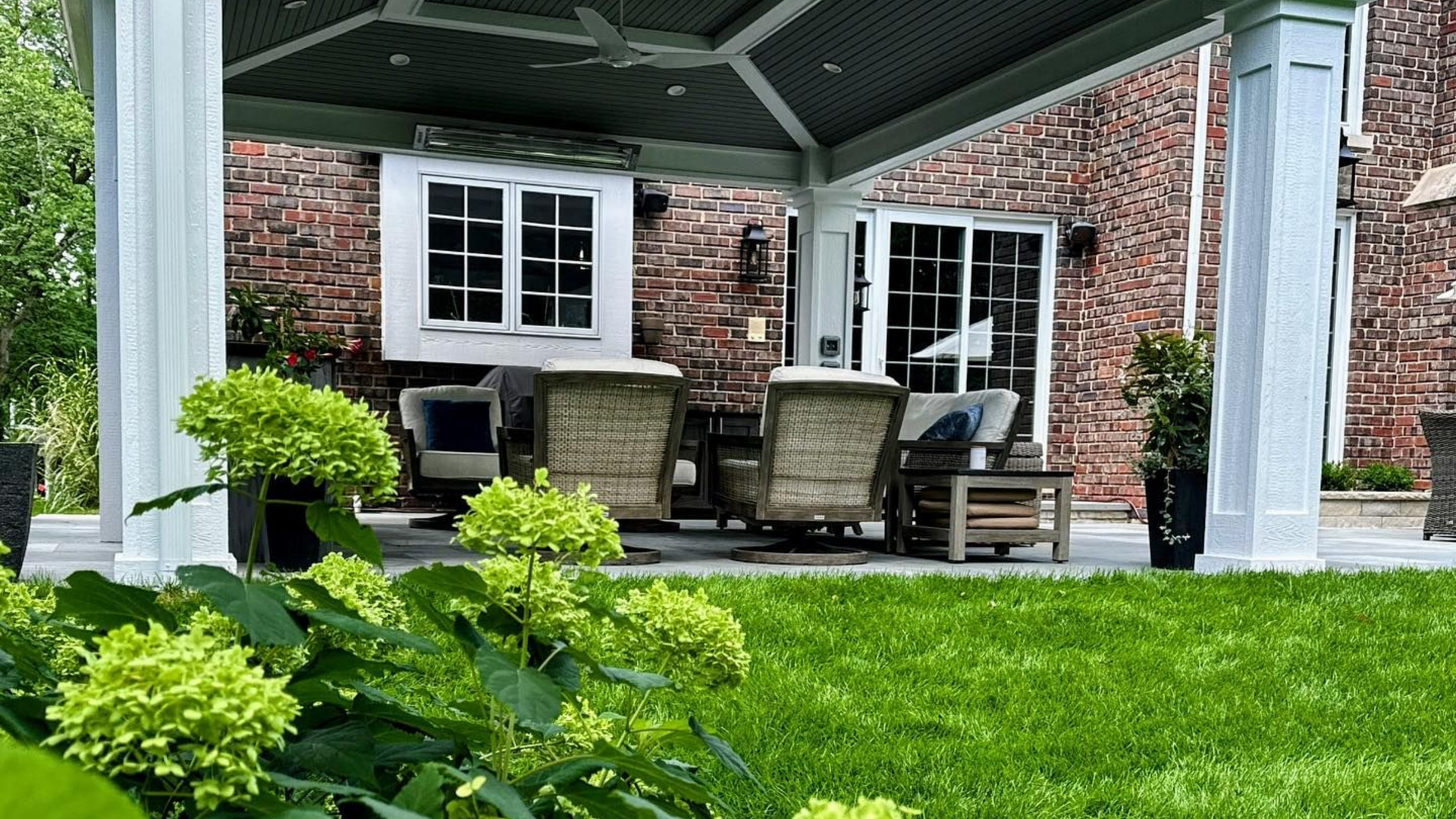 Covered patio with wicker furniture and brick wall backdrop, surrounded by greenery.
