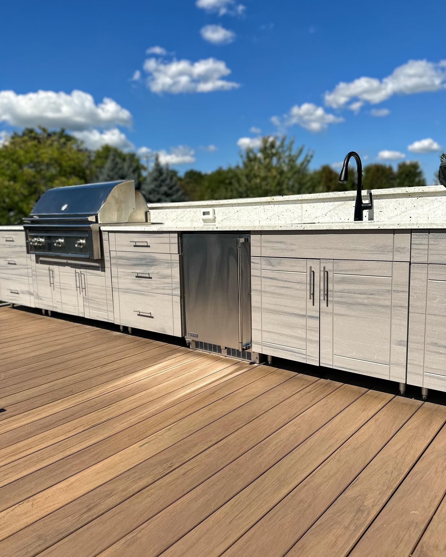 Outdoor kitchen with a grill, refrigerator, sink, and cabinetry on a wood deck under a blue sky.