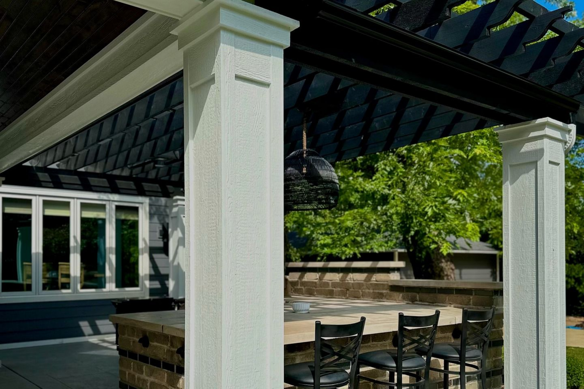 Outdoor kitchen with a pergola, bar seating, and stone structure, with a view of a house and trees.