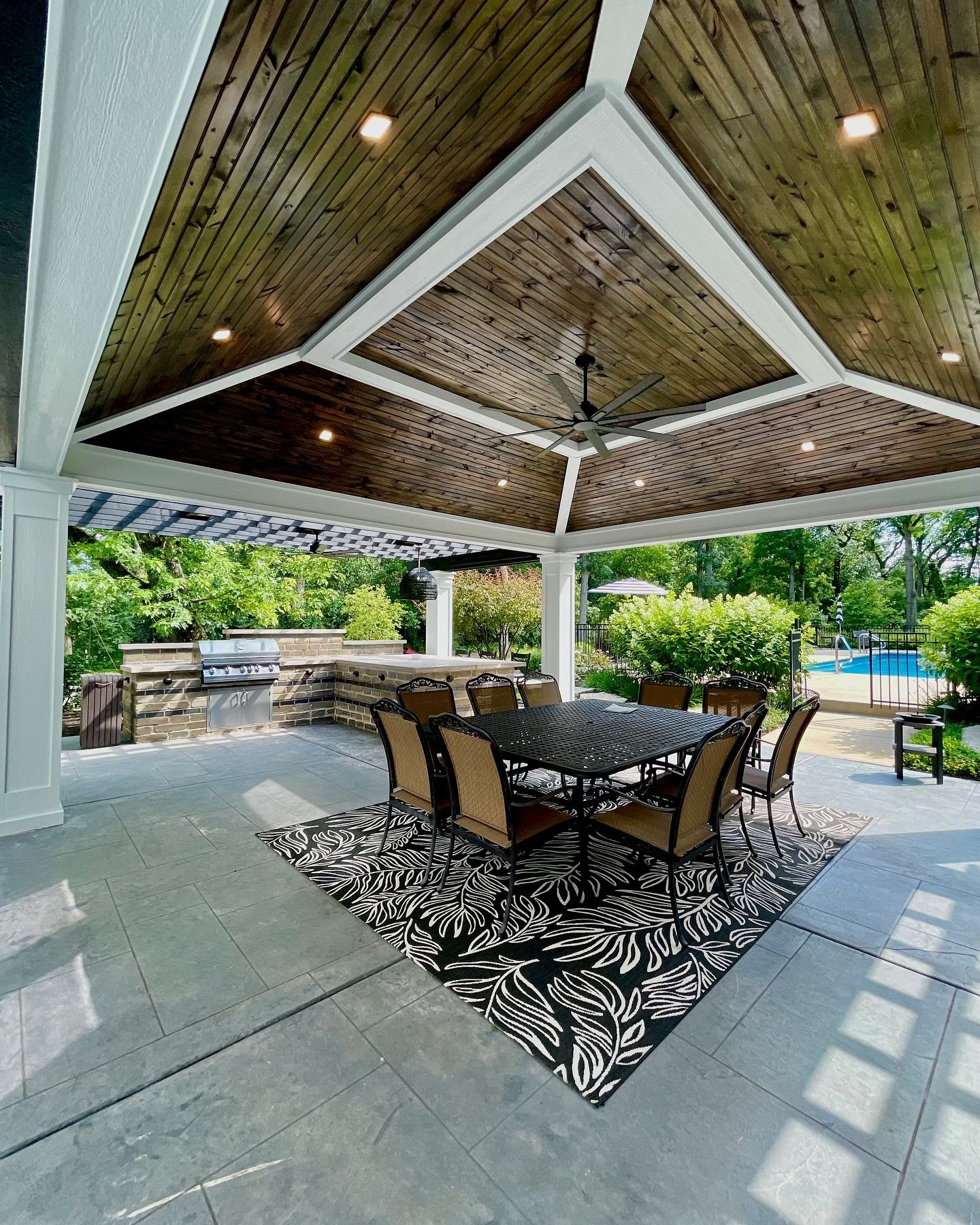 Patio with wooden ceiling, dining set on a rug, pool in background.