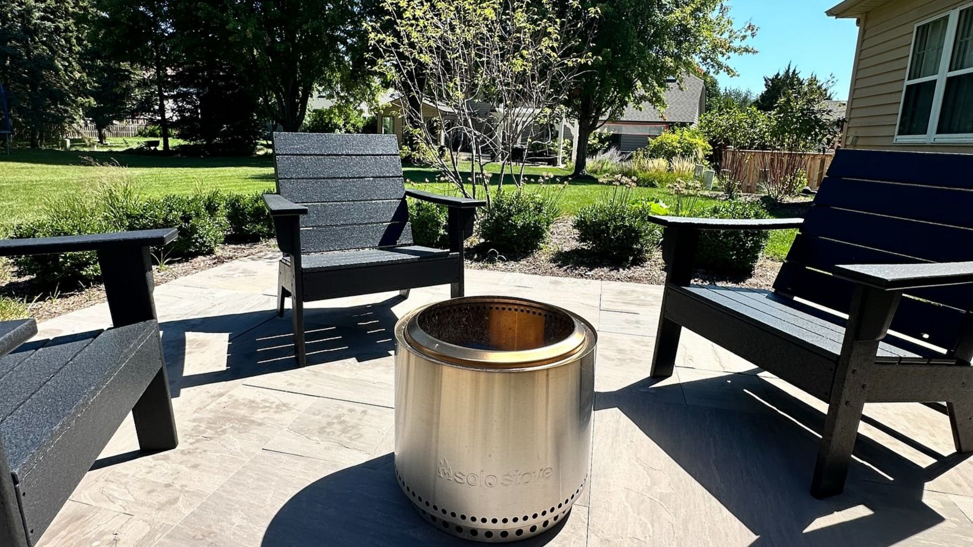 Outdoor patio with a fire pit, three black chairs, and trees in the background on a sunny day.