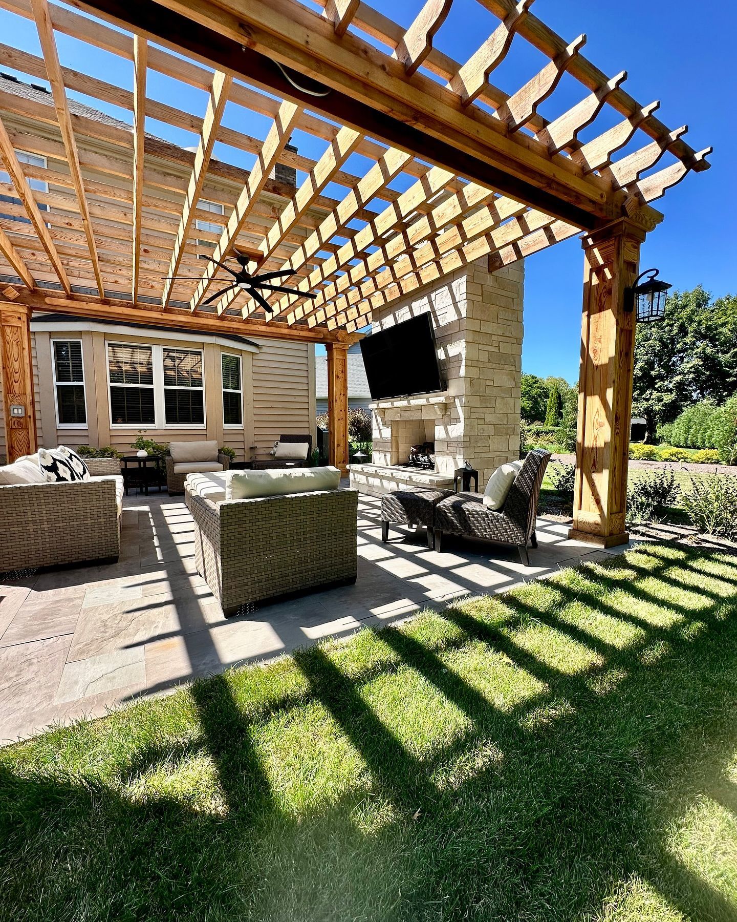 Wooden pergola over patio with seating, fireplace, and TV; bright sunlight casts shadows.