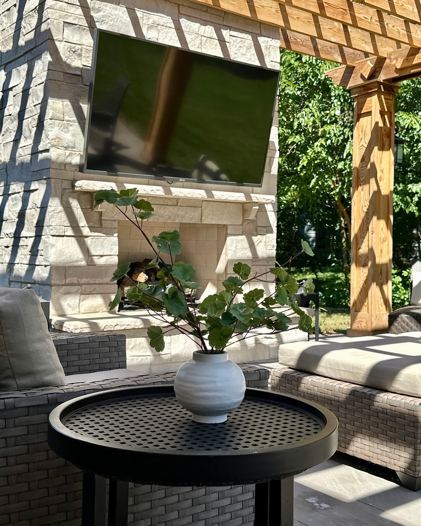 Outdoor patio with a TV above a fireplace, table with vase, and seating under a pergola.