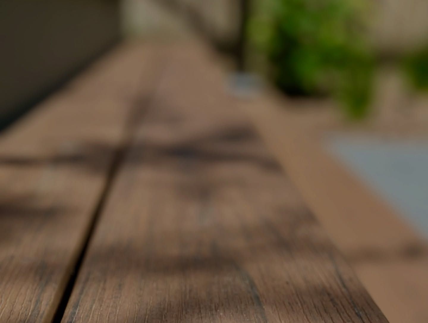 Close-up of brown wooden planks, with blurred background of a patio and green foliage.