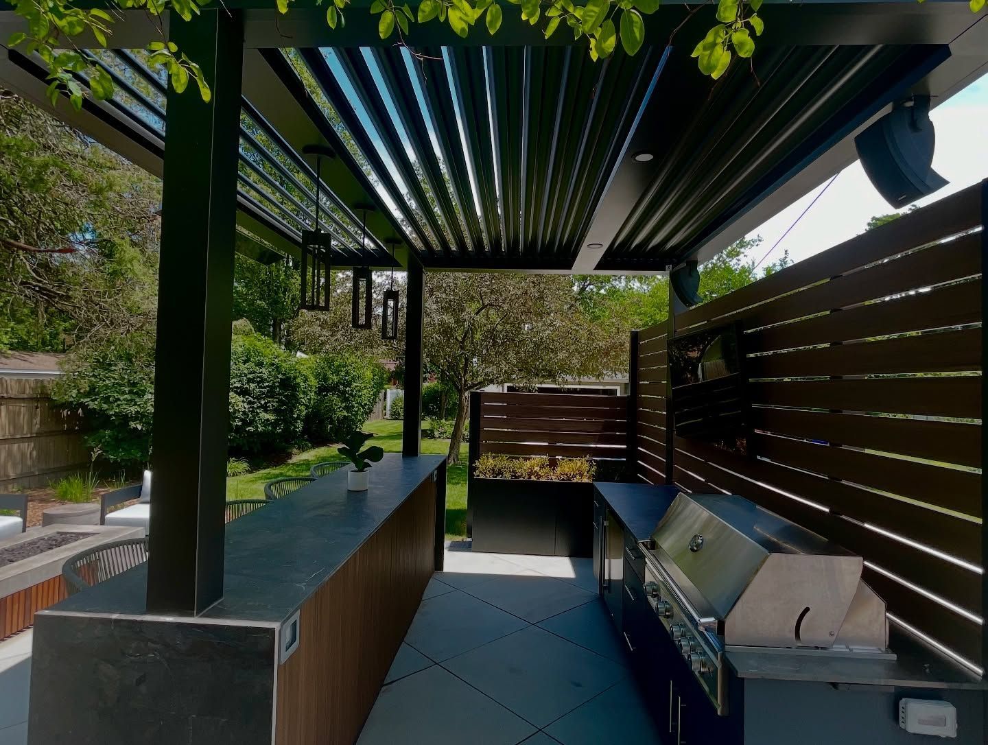 Outdoor kitchen under a modern pergola with grill, bar, and privacy fence.