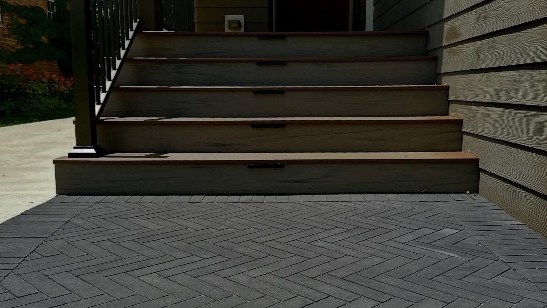 Gray brick herringbone pattern walkway leading to concrete steps with a black railing, and a red door.
