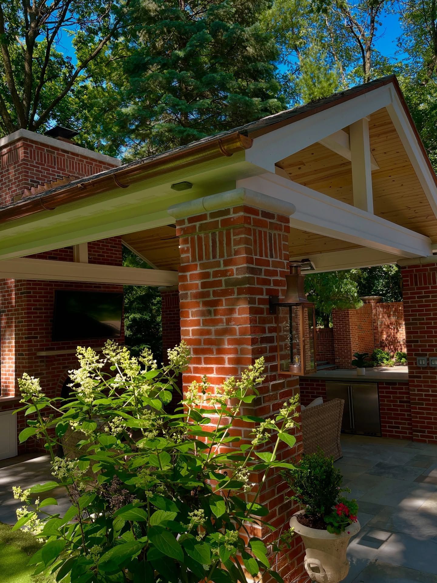 Red brick outdoor structure with wooden beams and a green canopy, set in a lush garden.