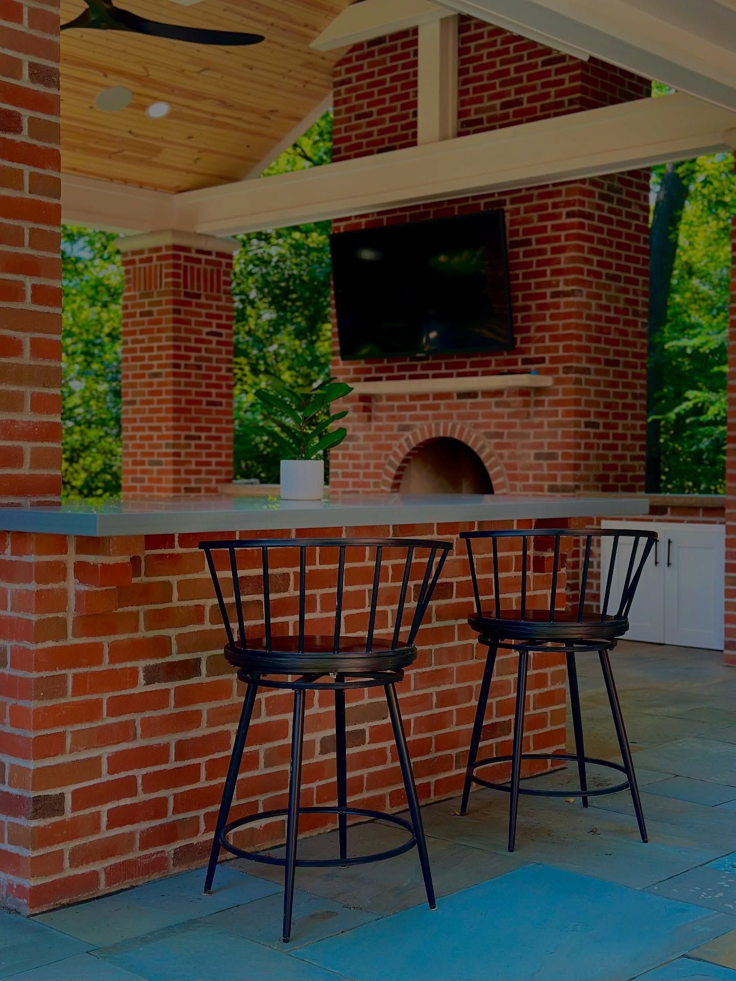 Outdoor bar with brick facade, stools, and a fireplace with a TV. Lush greenery in the background.
