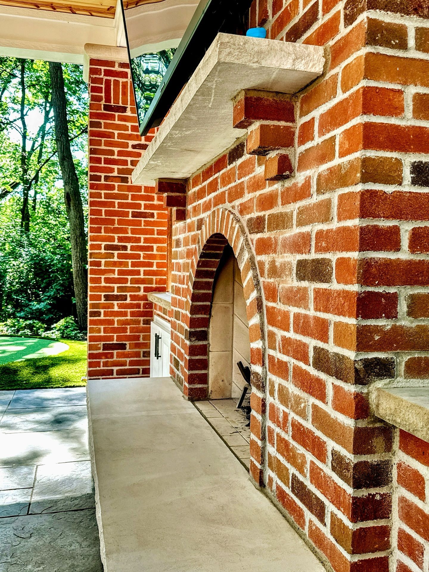Brick outdoor fireplace with arched opening, concrete shelf, and green trees in background.