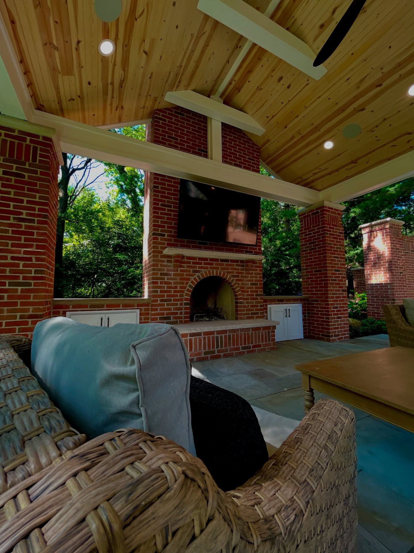 Outdoor brick fireplace and TV on a patio with wicker furniture, under a wood ceiling.