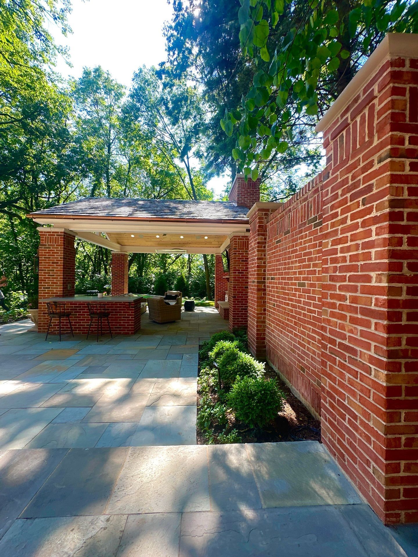 Brick patio with a covered seating area and wall, surrounded by trees.