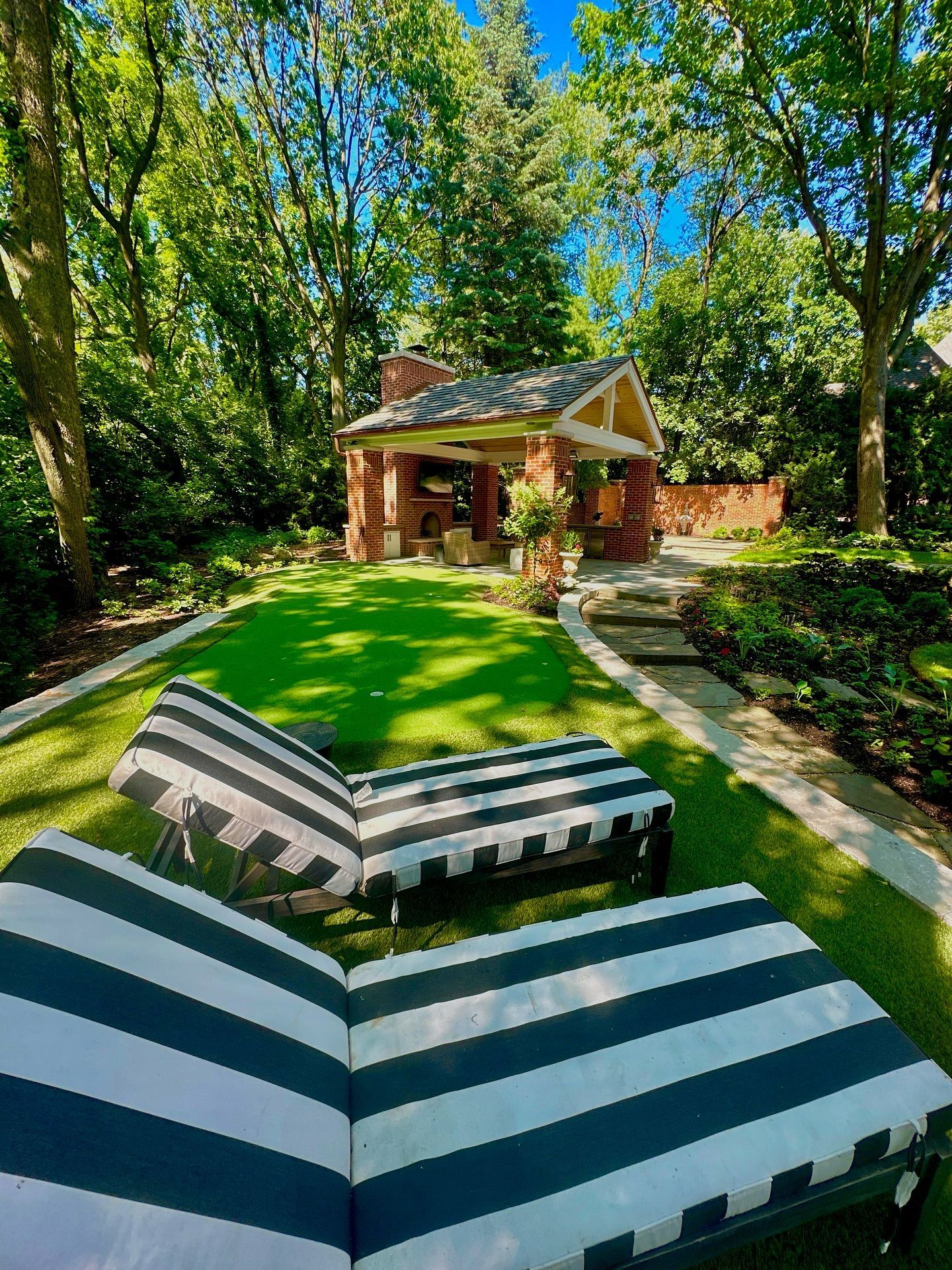 Two striped lounge chairs on green lawn, brick gazebo in background, surrounded by trees.