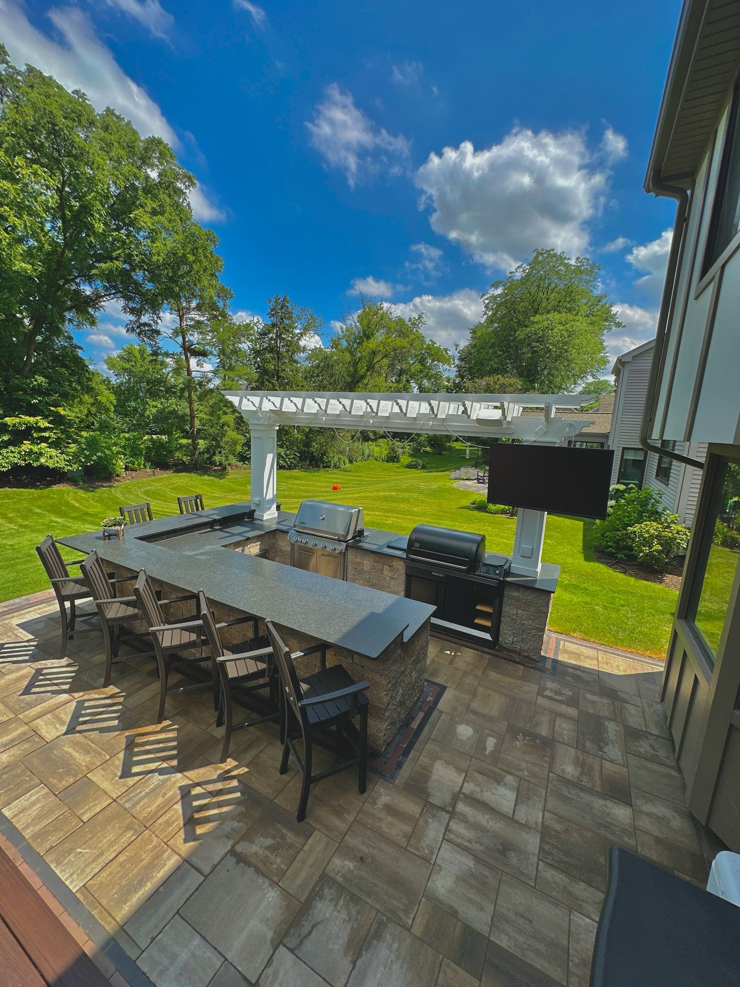 Outdoor kitchen with a long countertop, grill, and TV on a patio with a pergola; sunny day.
