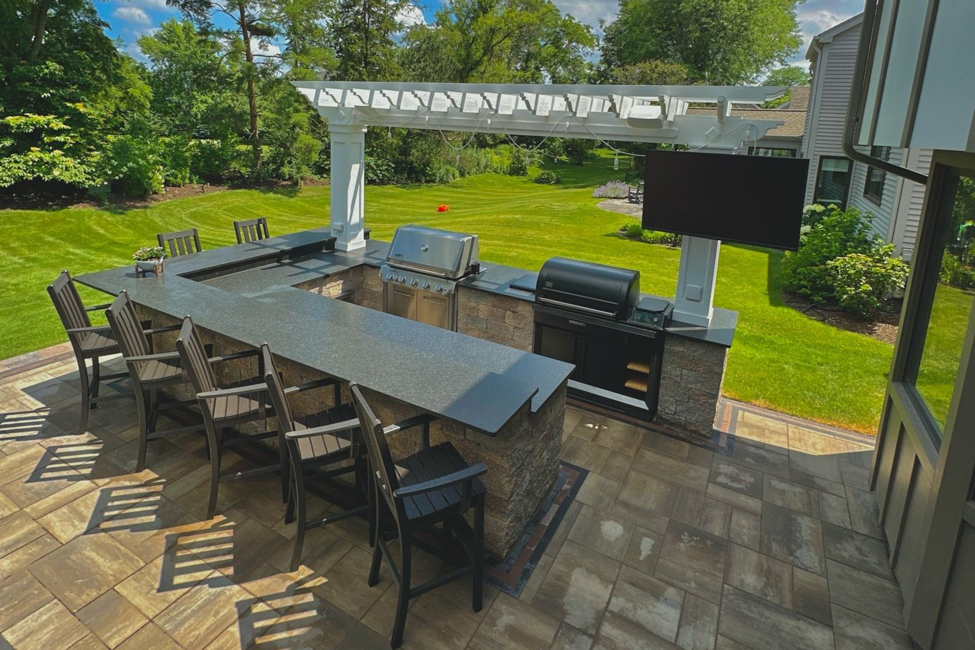Outdoor kitchen with a bar, grill, and a TV under a pergola on a stone patio.
