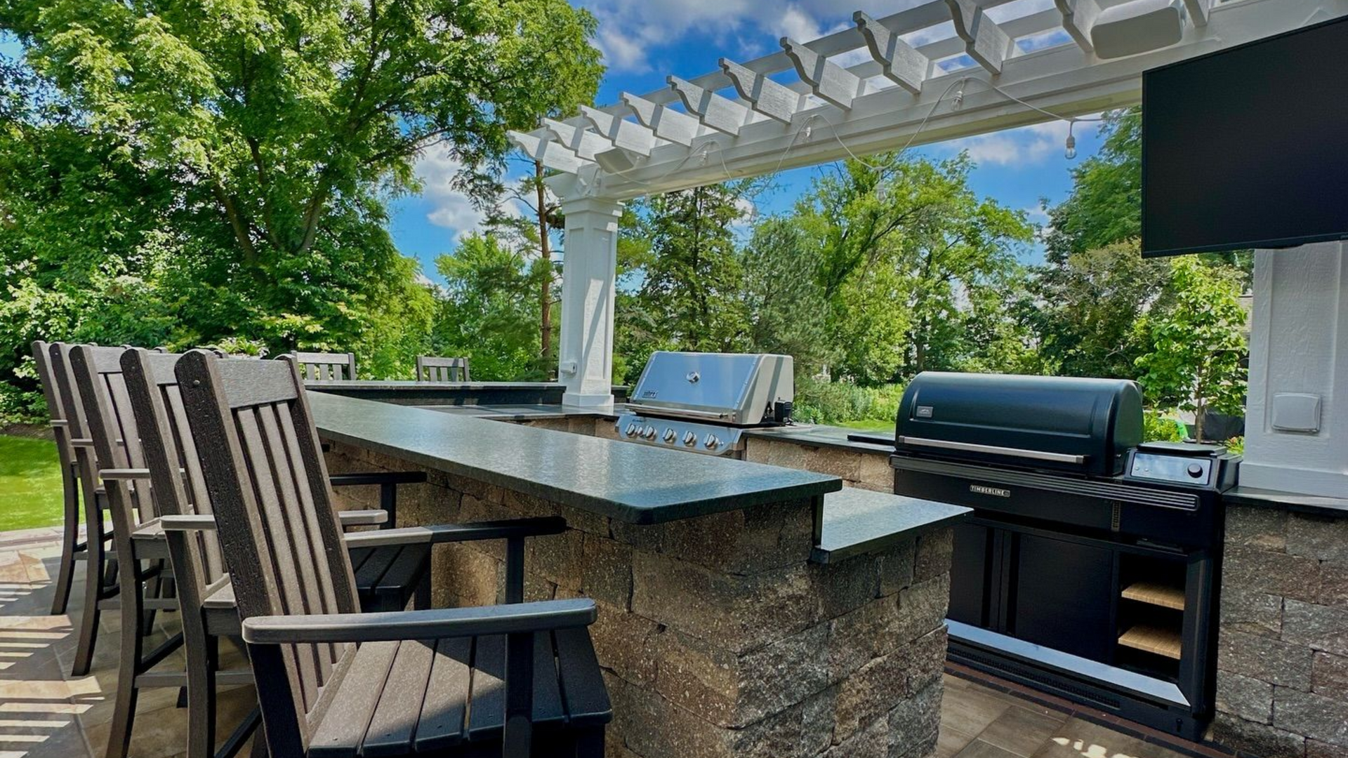 Outdoor kitchen with a grill, countertop, bar stools, and a pergola.