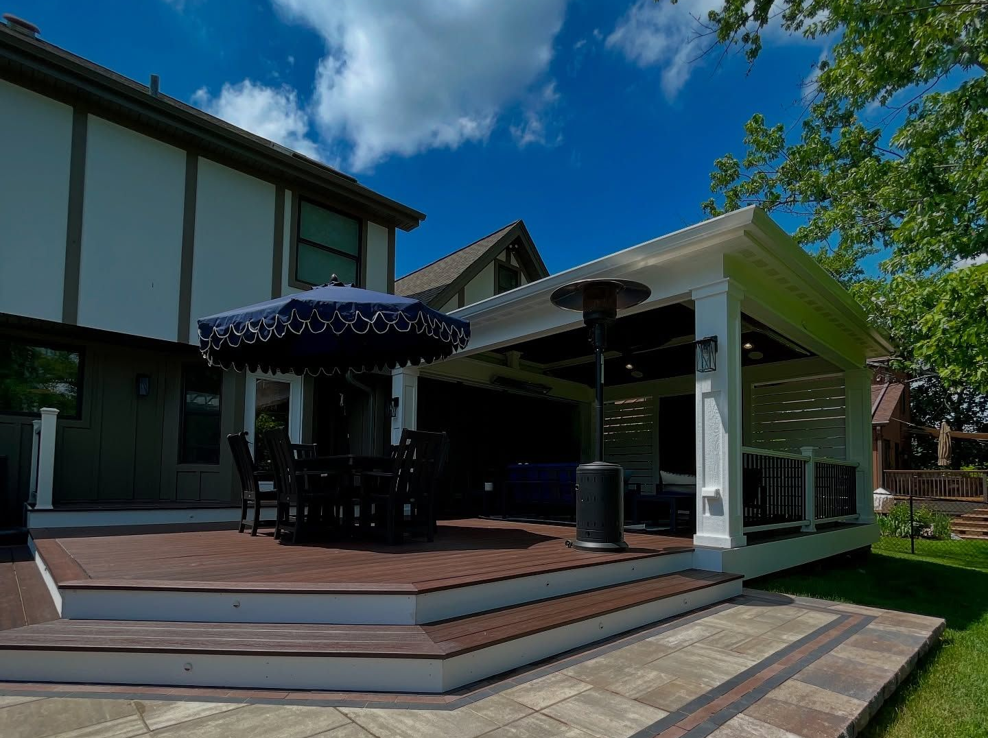 Backyard patio with deck, awning, seating, and pavers on a sunny day.