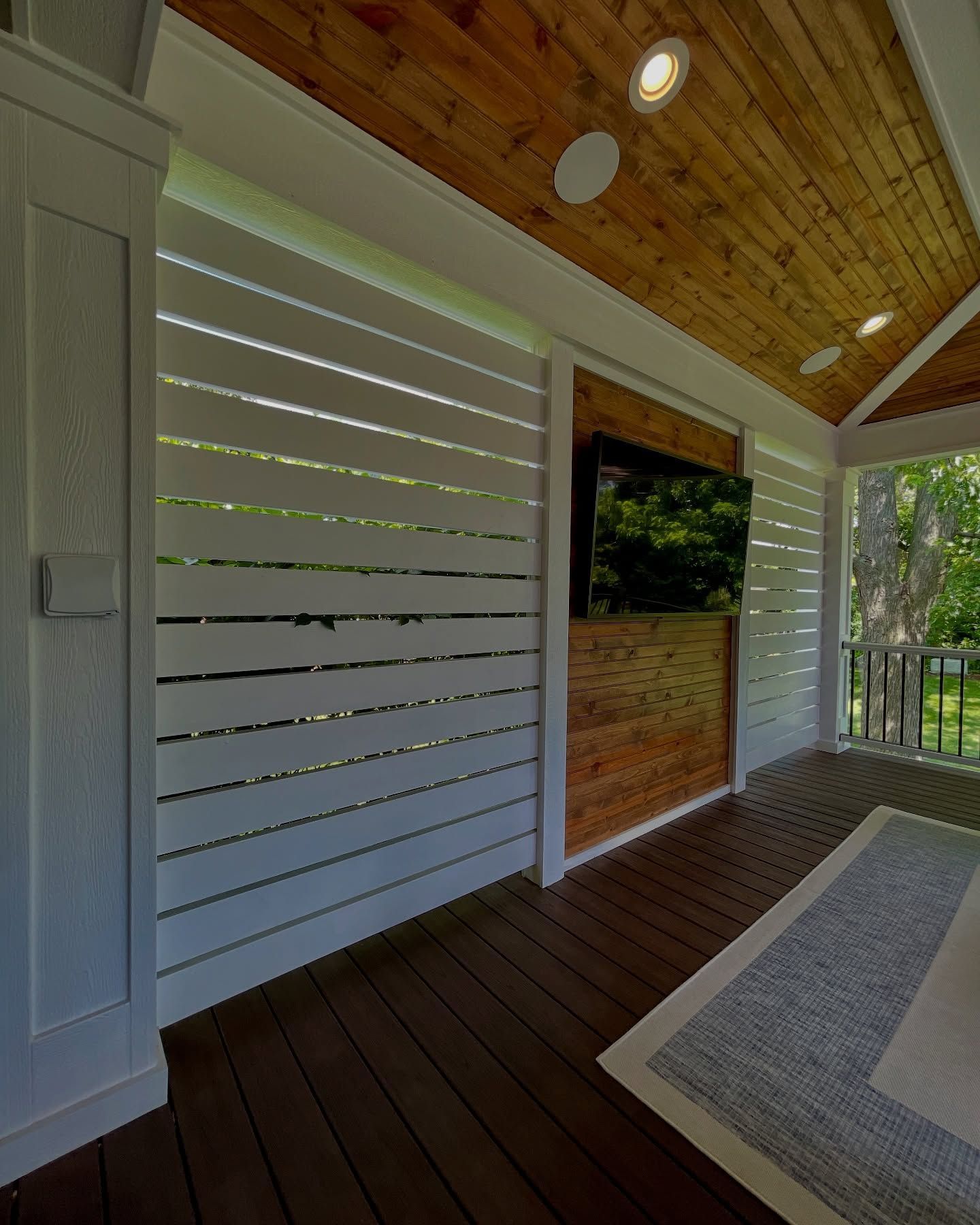 Outdoor covered porch with white slat wall, wood ceiling, and mounted TV.