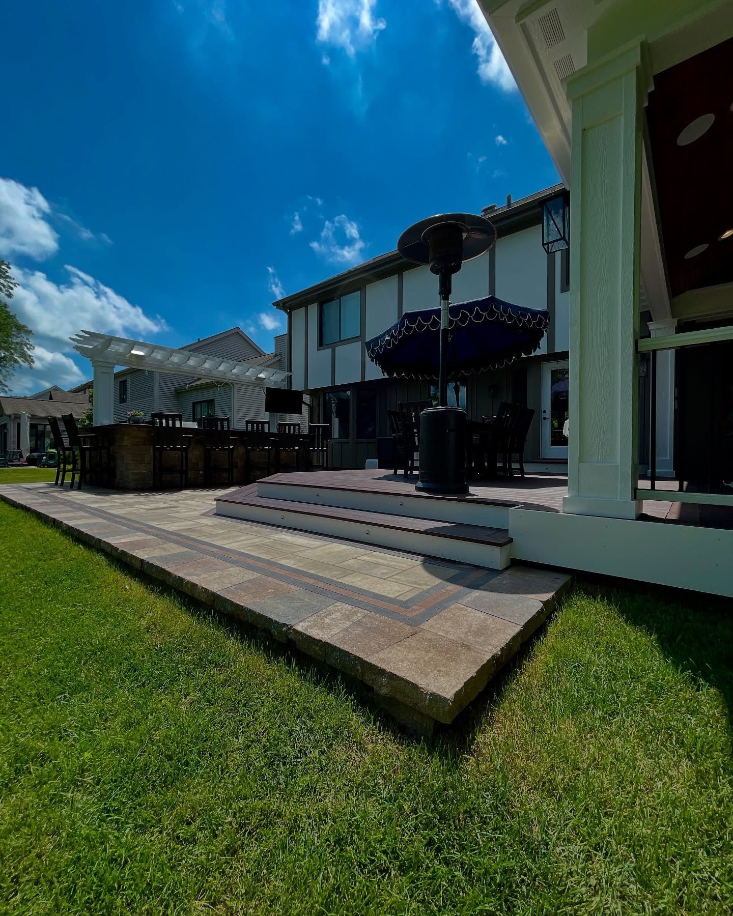 Backyard patio with brick steps, outdoor kitchen, and dining set under a bright blue sky.