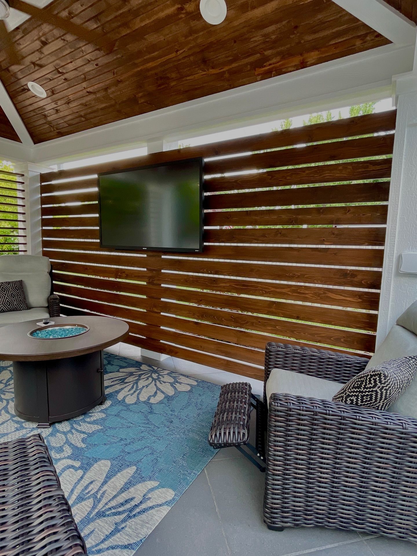 Outdoor seating area with wood-slatted wall, mounted TV, rug, fire pit table, and woven furniture.