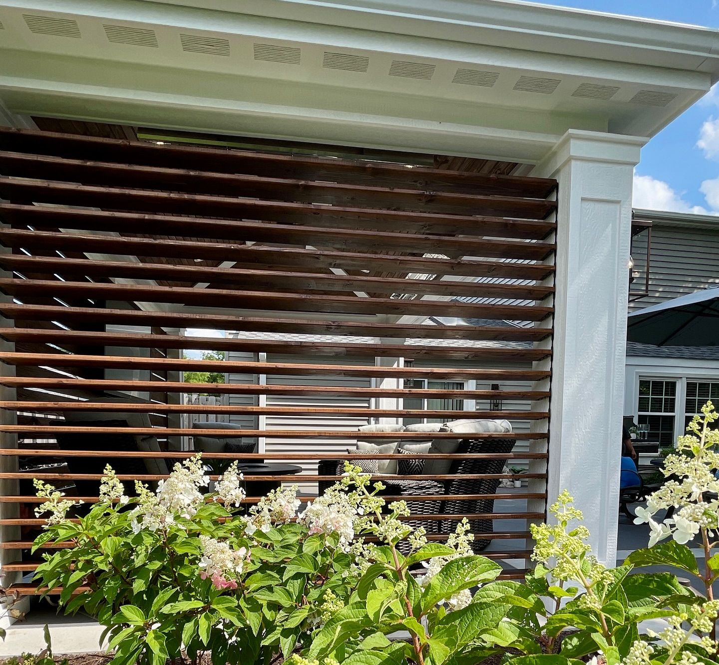 Brown horizontal slats screen a building facade with white columns; flowers in front.