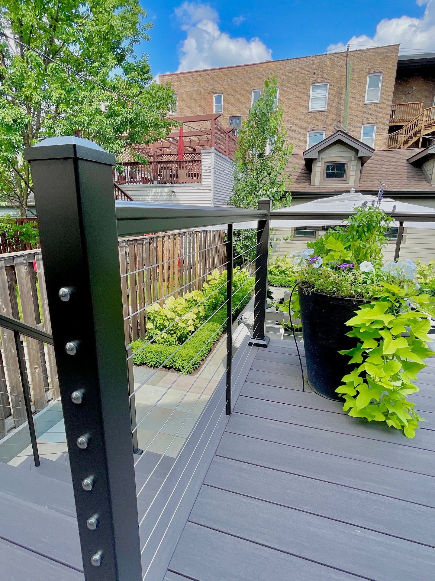 A rooftop deck with gray flooring, black railing, and potted plants, overlooking a building and trees under a blue sky.
