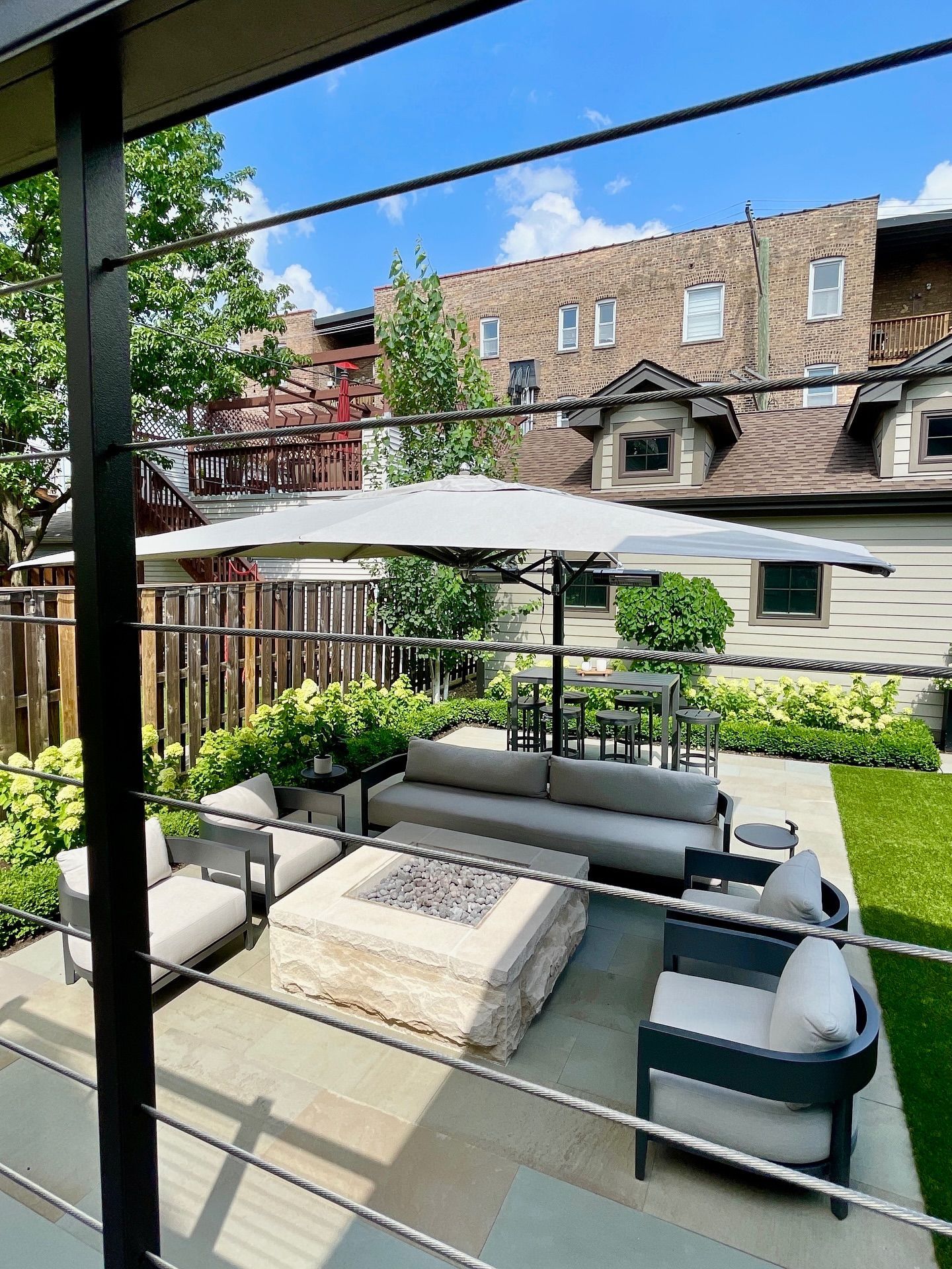 Patio with outdoor furniture, fire pit, and umbrella. Green lawn and a brick building in the background.