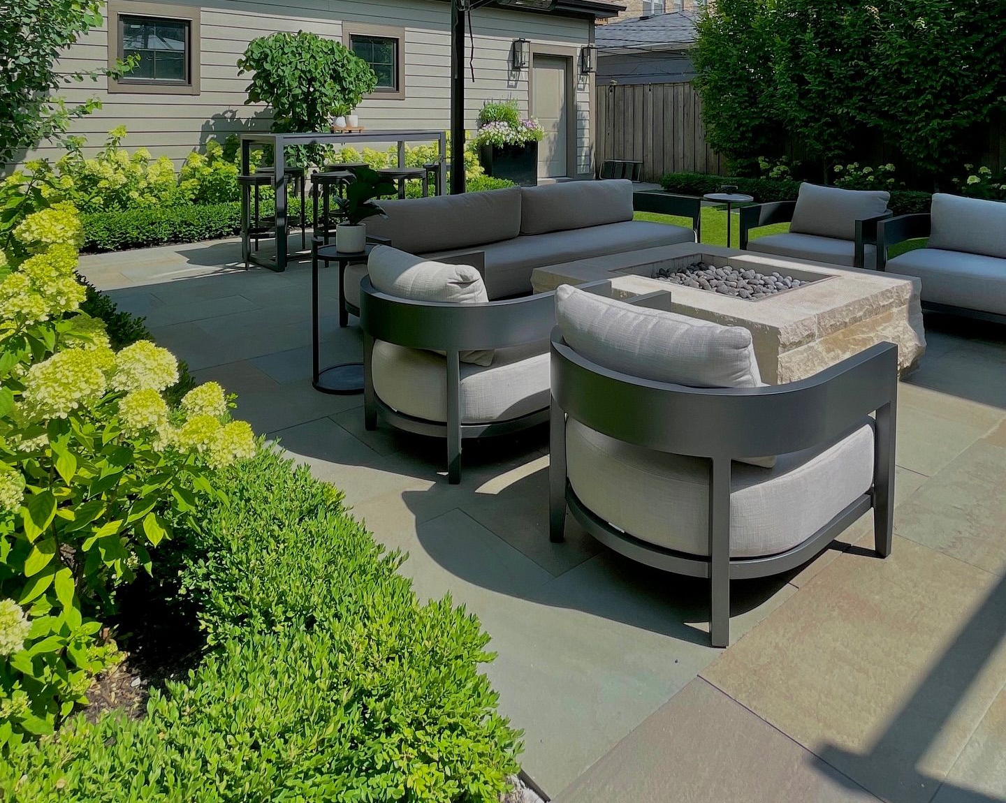 Patio with modern gray furniture, fire pit, and lush green plants.