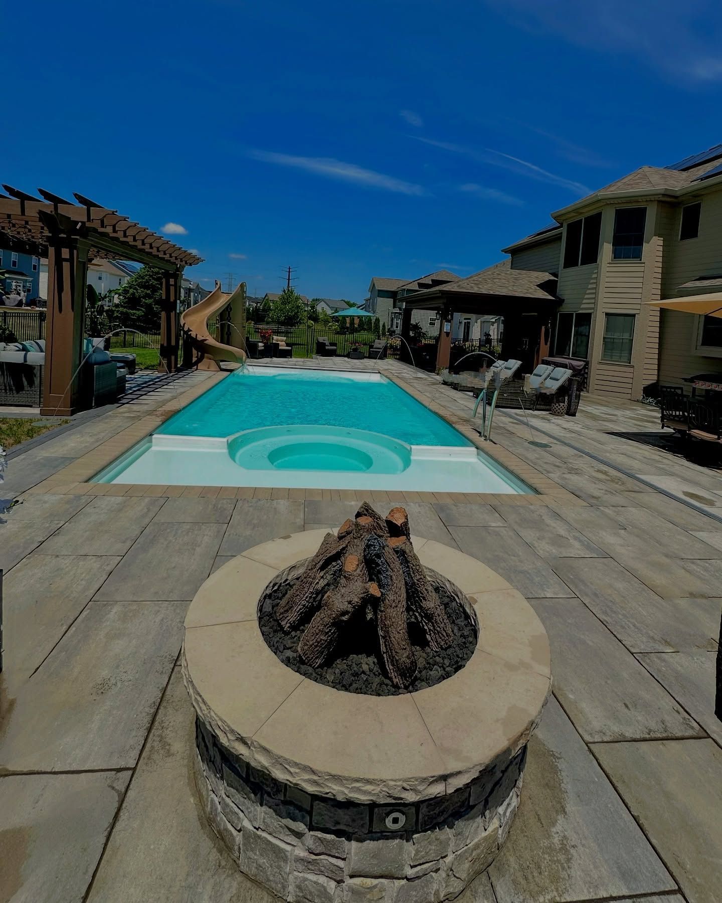 Backyard with pool, jacuzzi, fire pit, and houses under a bright blue sky.
