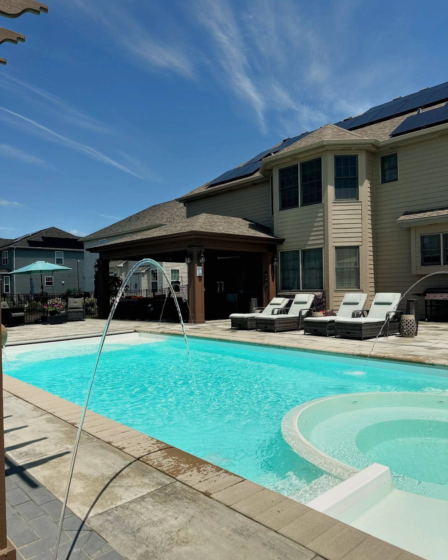 Swimming pool with fountain in front of a house, gazebo, and lounge chairs. Sunny, blue sky.