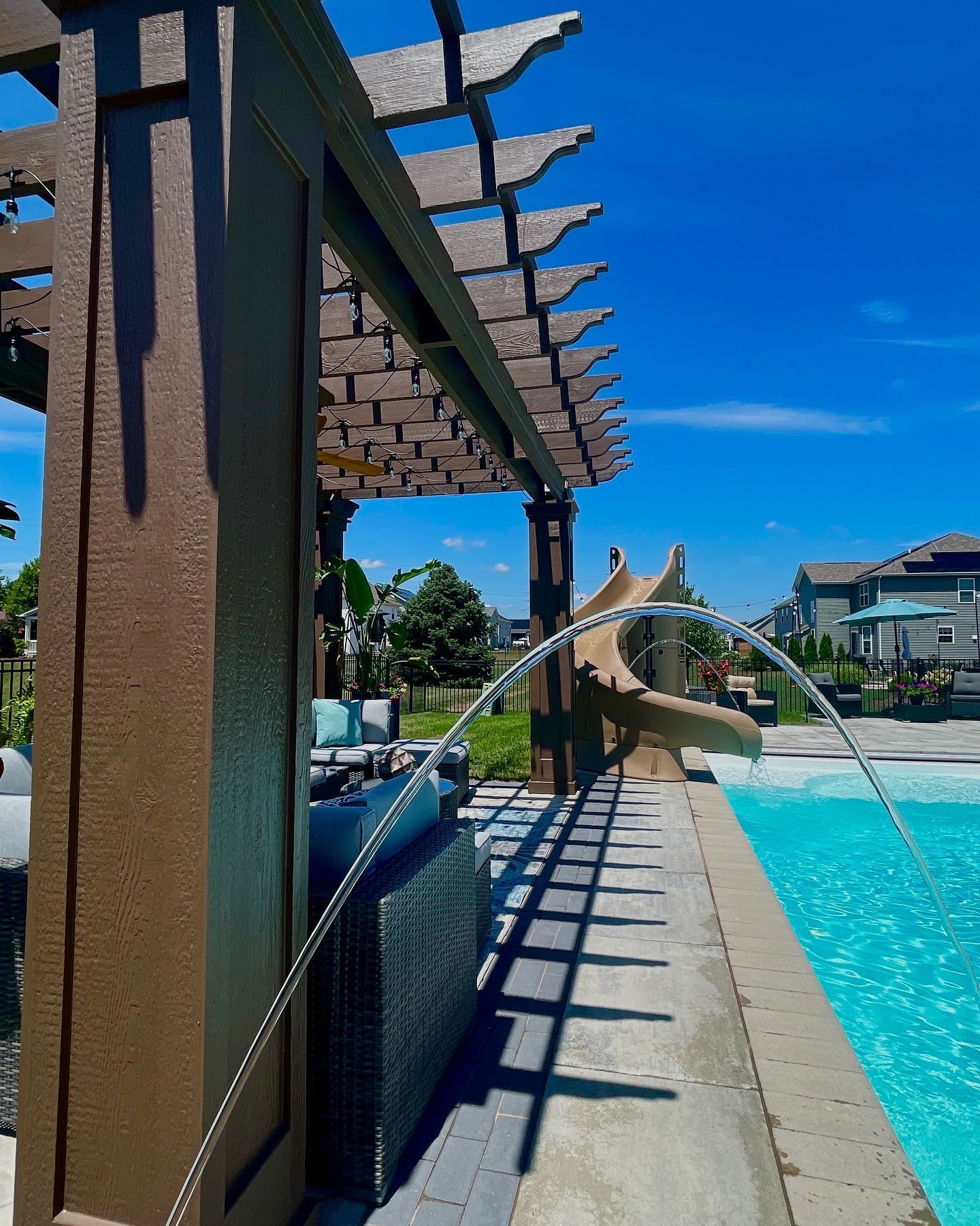 Brown pergola next to pool, water fountain, blue sky, and a water slide.