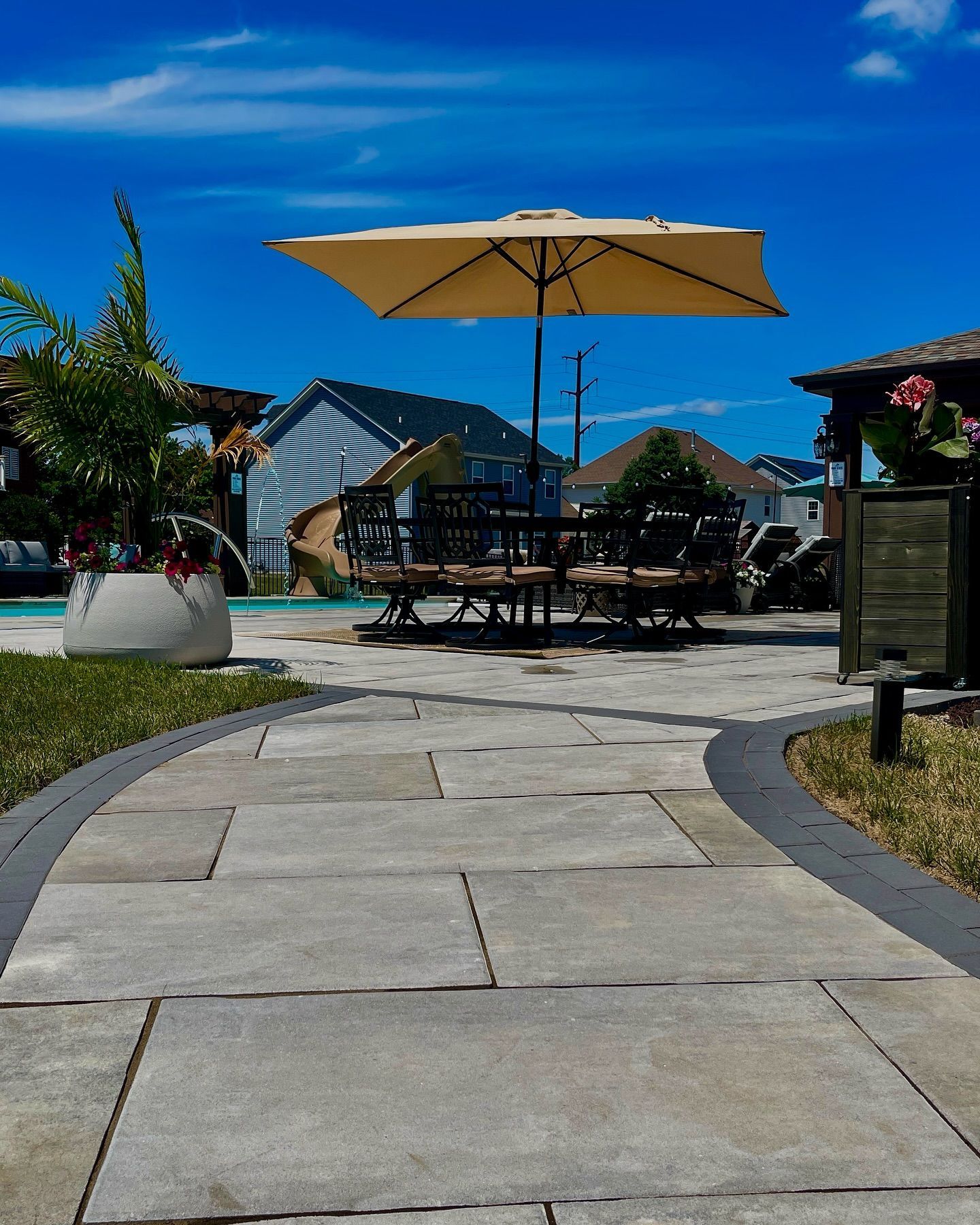 Stone pathway leading to an outdoor dining area with a beige umbrella and tables under a blue sky.