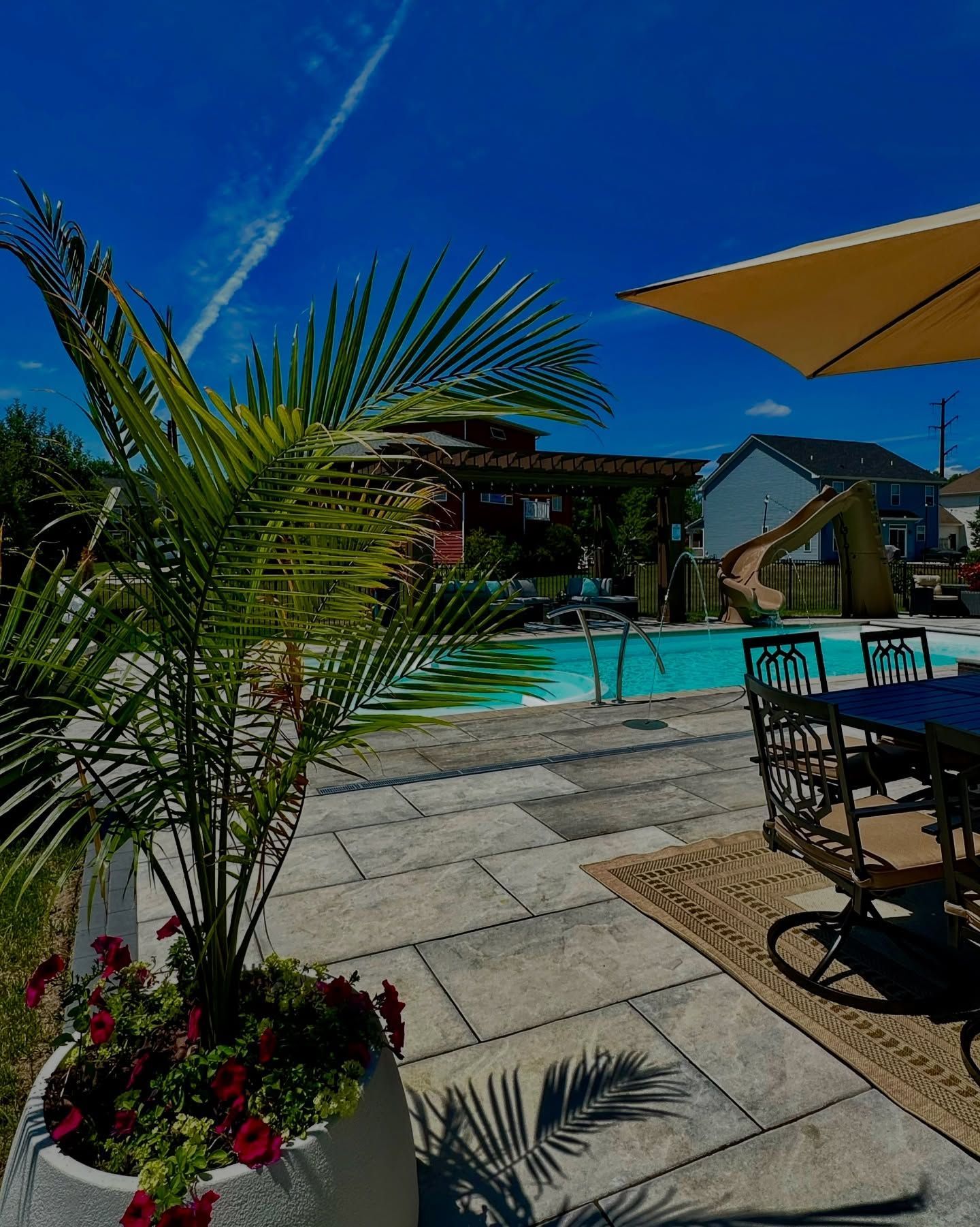 Poolside scene with a palm tree, dining table, blue sky, and a swimming pool.