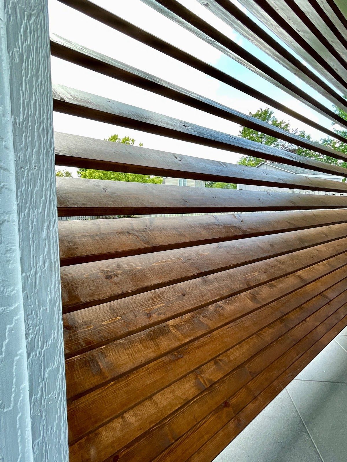 Wooden slats on a wall, filtering sunlight, with a glimpse of greenery and sky.