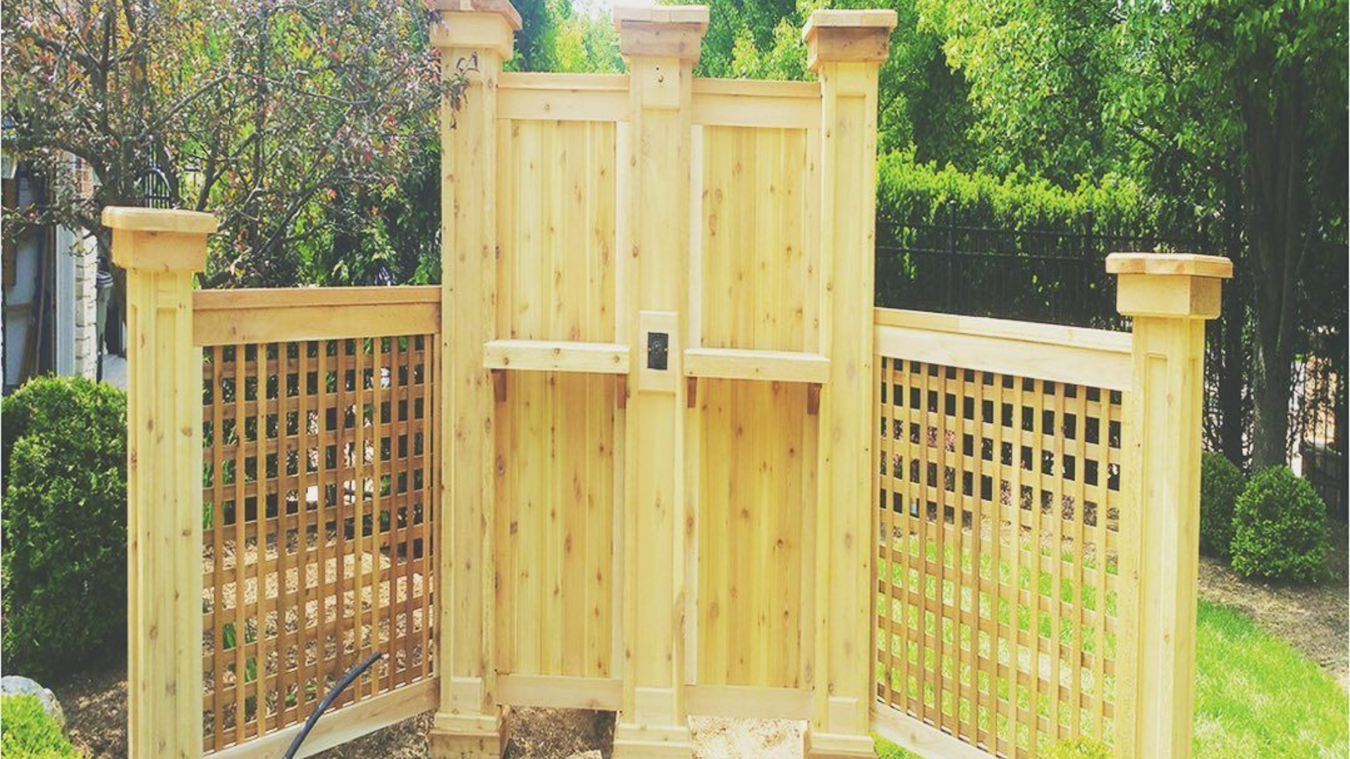 Wooden outdoor shower with lattice side panels and decorative posts, surrounded by greenery.