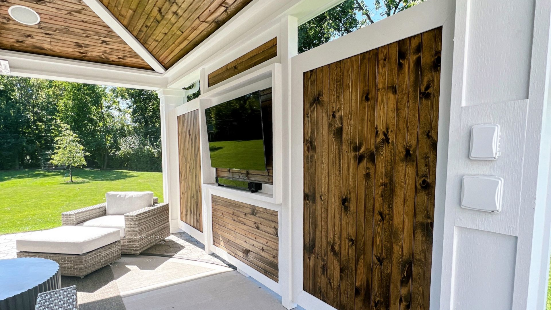 Covered patio with wood-paneled walls, outdoor TV, and wicker seating, set against a green lawn.
